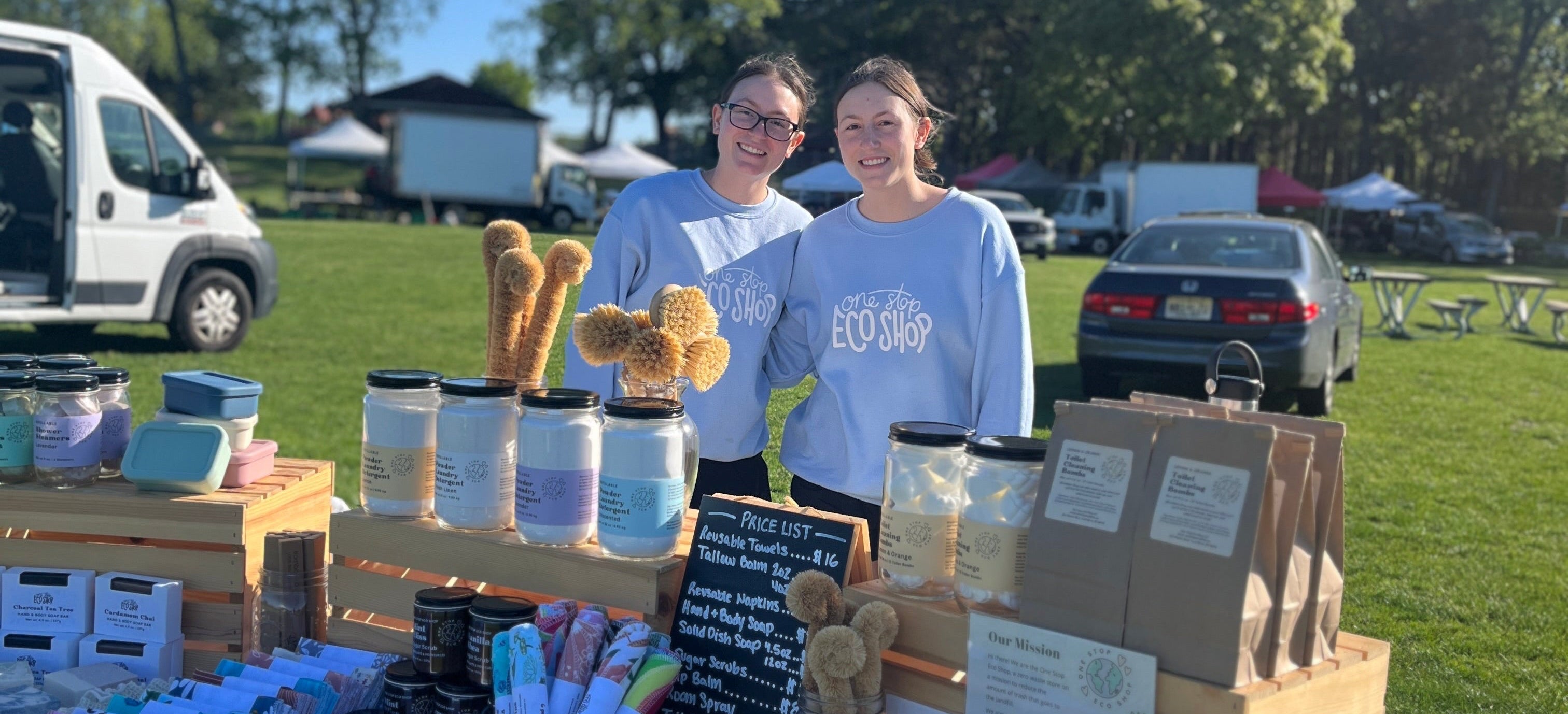 Two women standing behind a table with various items at an outdoor market.